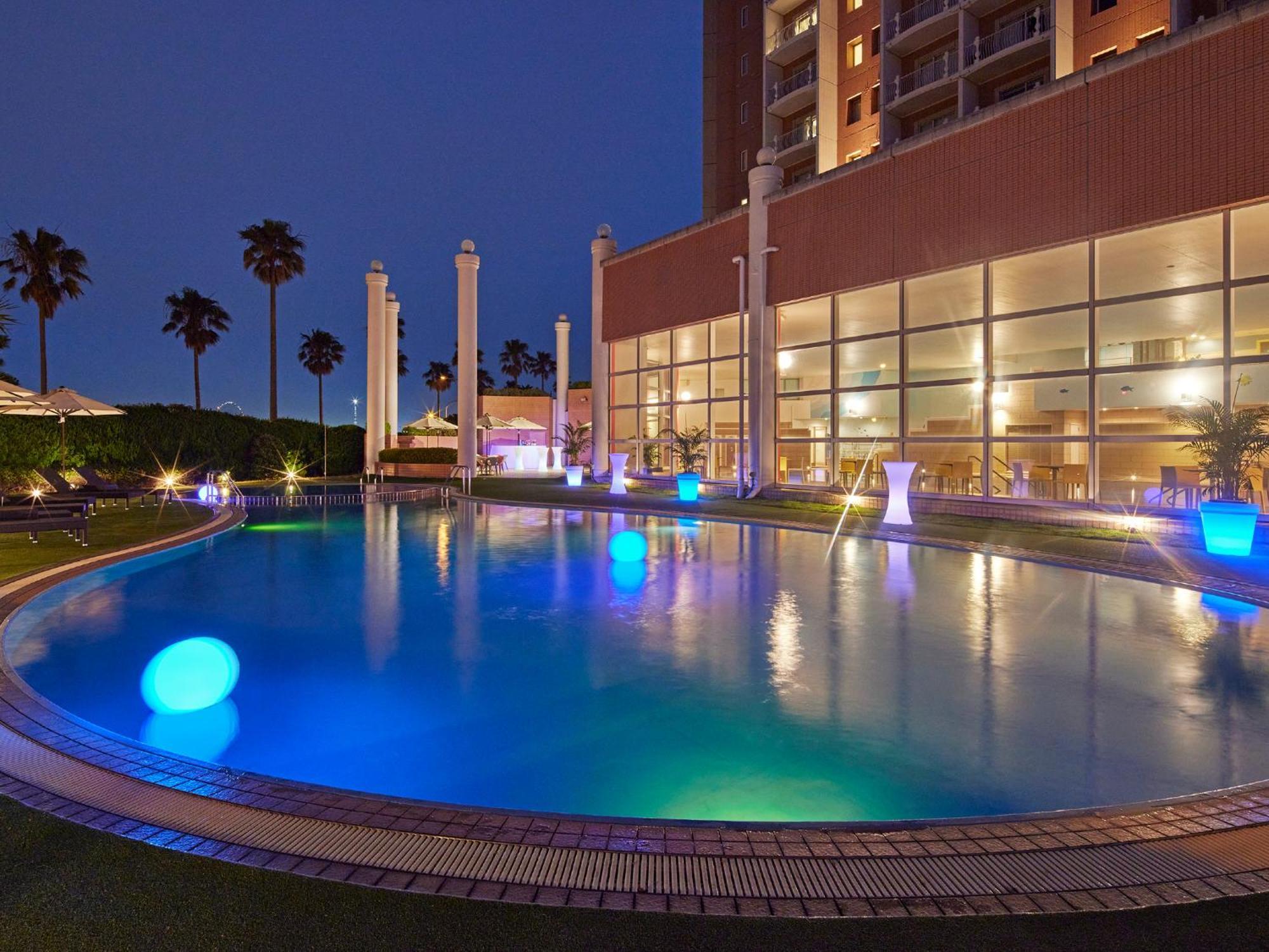 Grand Nikko Tokyo Bay Maihama Hotel Urayasu Ngoại thất bức ảnh The photo shows a well-lit swimming pool at night, surrounded by a lush green area. The pool features glowing blue lights that create a serene and inviting atmosphere. Palm trees can be seen in the background, adding a tropical feel to the scene. Add